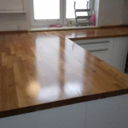 Kuchenny blat drewniany z widocznymi słojami, połączony z białymi szafkami, odbijający światło okna. Ujęcie z bliska eksponuje strukturę drewna i wykończenie.