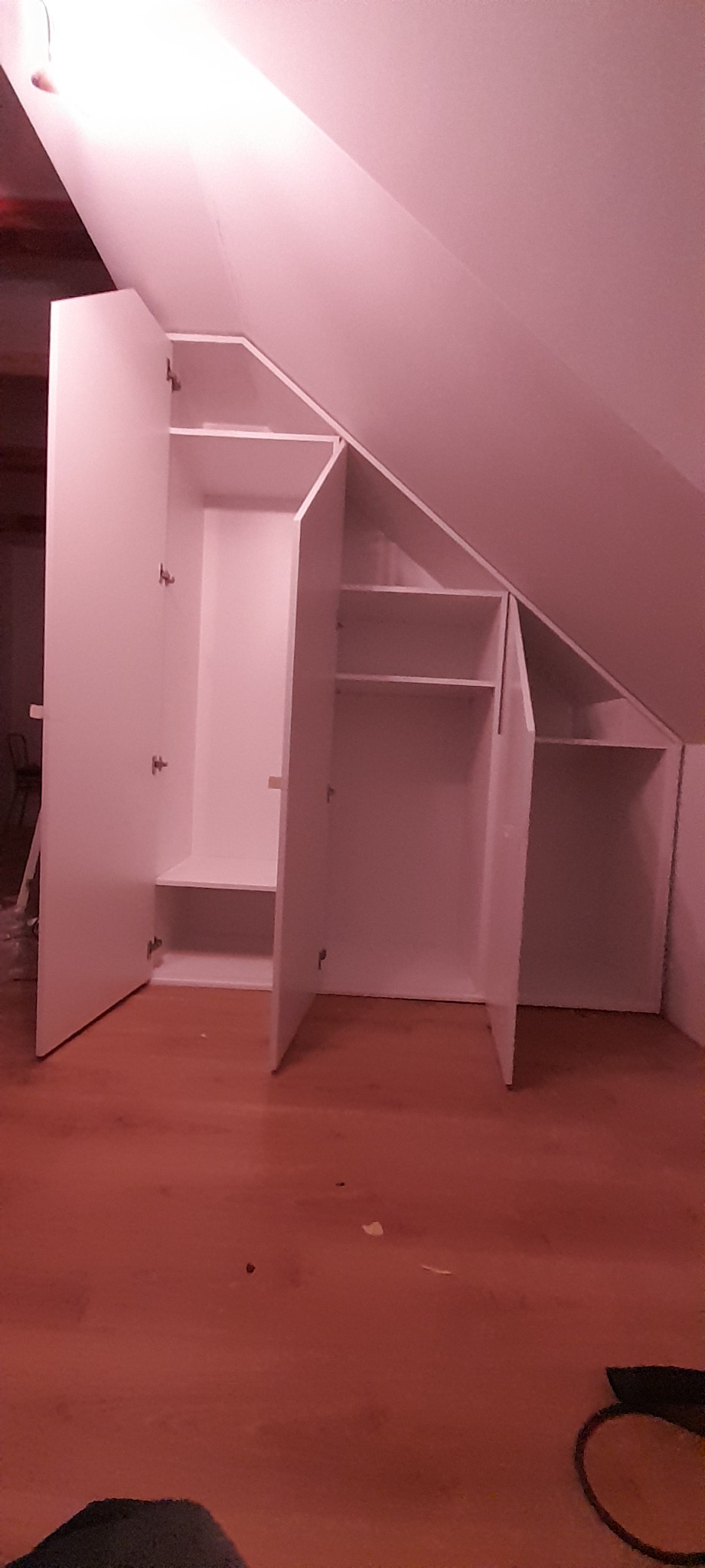 Biała szafa wnękowa na poddaszu, z otwartymi drzwiami, ukazująca półki i przestrzeń do wieszania ubrań. Widok z dołu, podkreślający dopasowanie do skosu dachu.