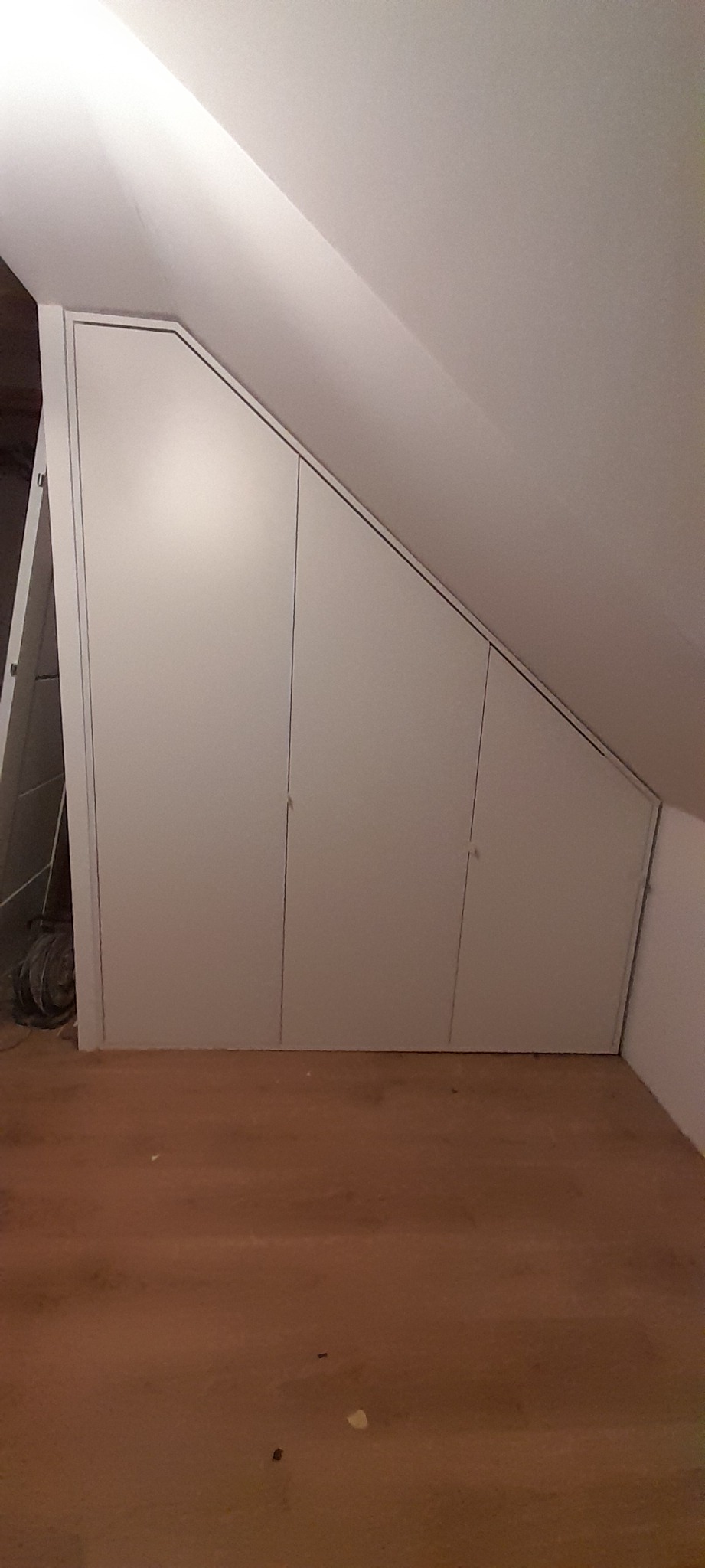 Biała szafa wnękowa na poddaszu, trzy skrzydła dopasowane do skosu dachu, drewniana podłoga, widoczna drabina i przewody elektryczne po lewej stronie.