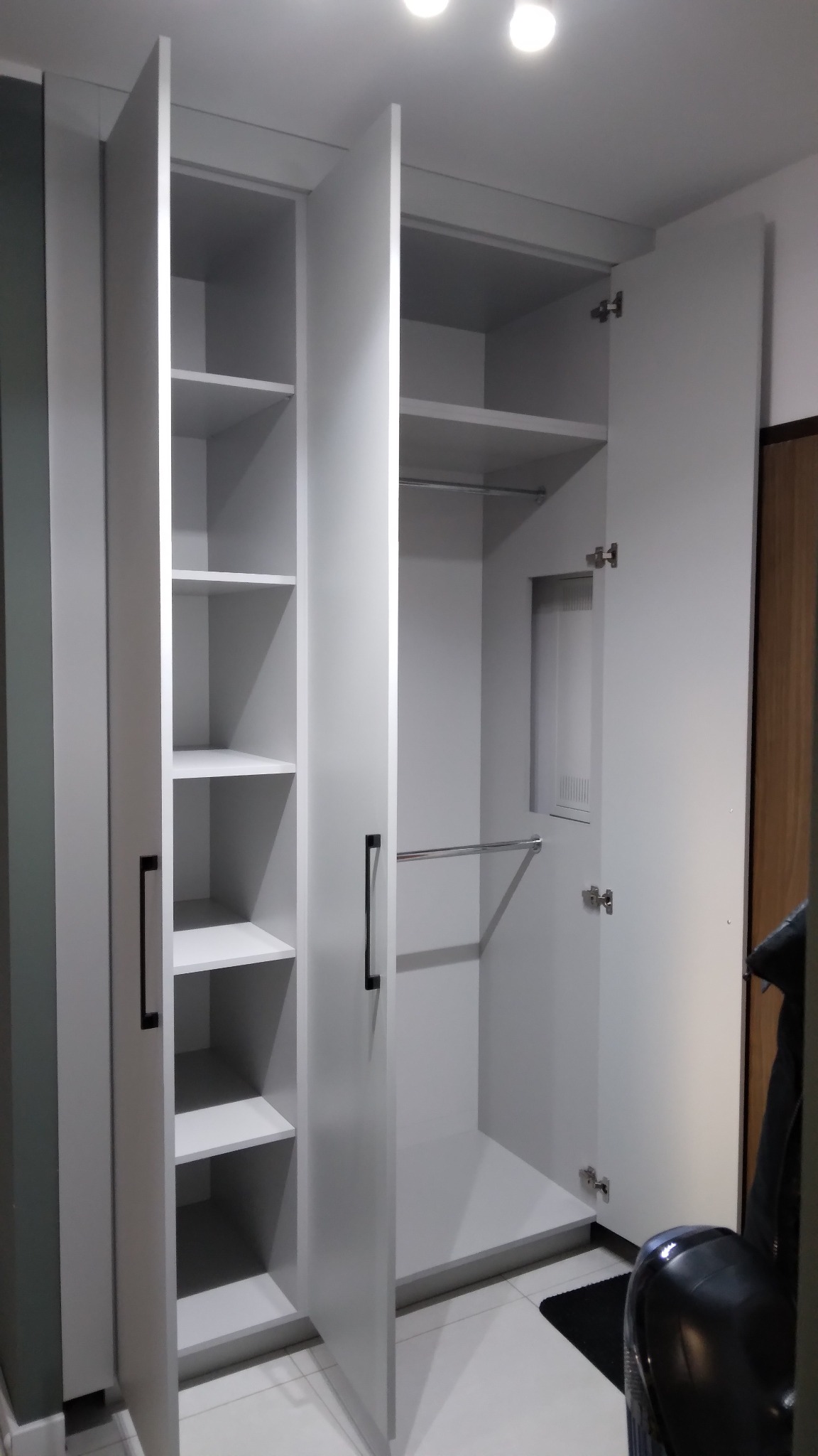 Otwarta, biała szafa wnękowa z półkami i drążkami na ubrania, widoczna na tle jasnej podłogi i ściany. Minimalistyczny design, czarne uchwyty.