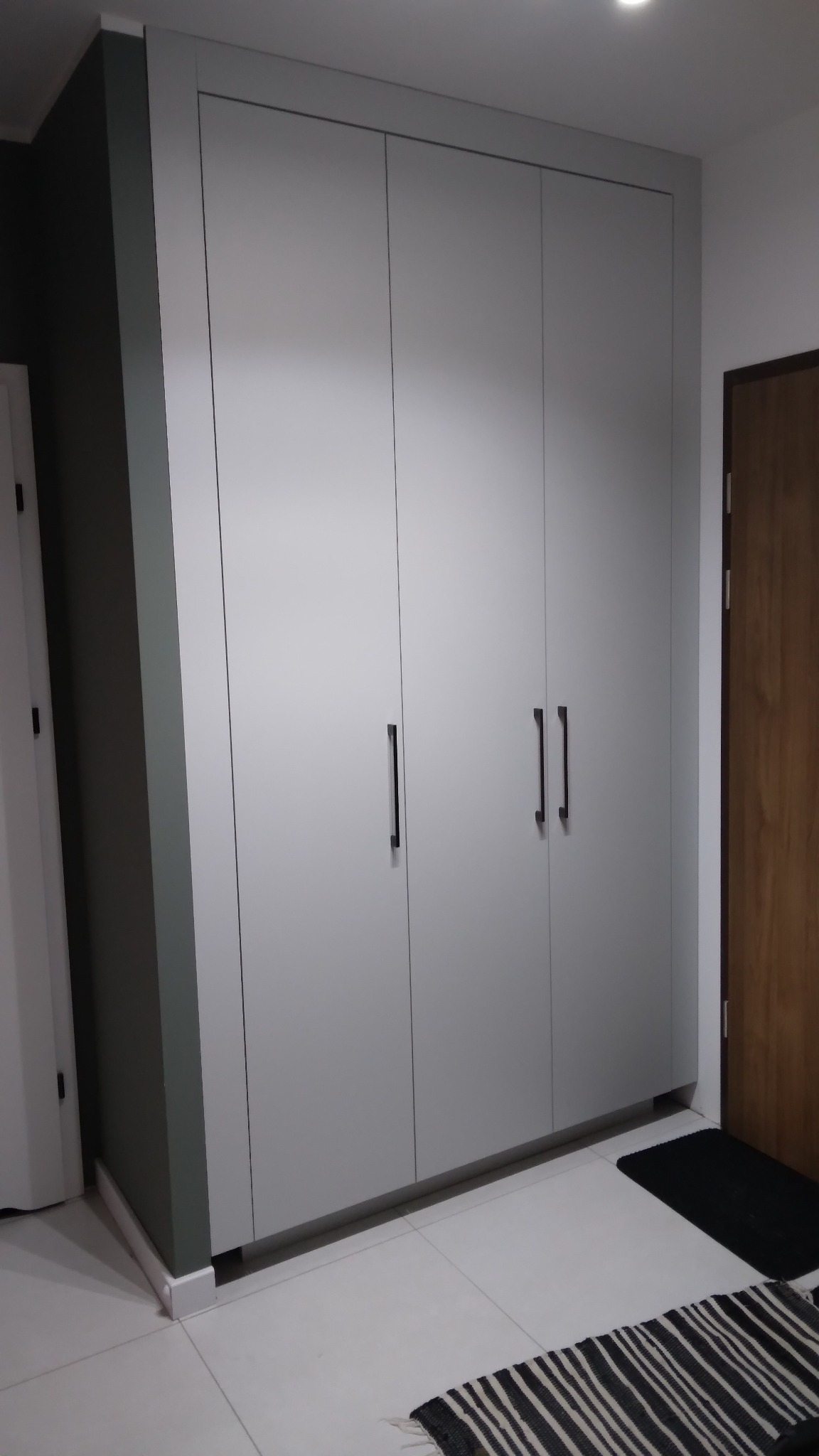 Czterodrzwiowa szafa wnękowa w odcieniach szarości z minimalistycznymi, czarnymi uchwytami, dopasowana do ściany. Widoczny fragment podłogi i dywanik.