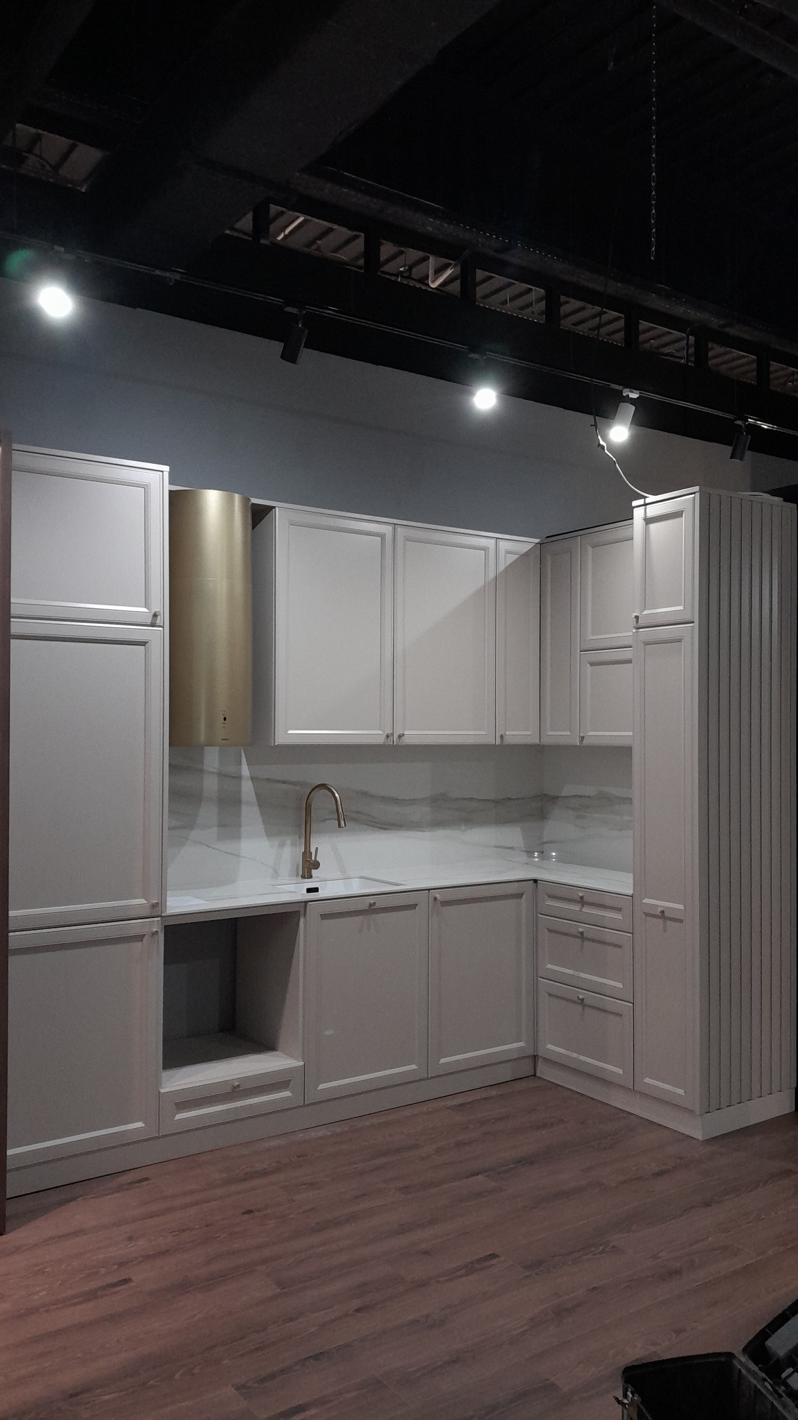 Jasna kuchnia z złotą armaturą i okapem na tle marmurowej ściany. Widoczne szafki górne i dolne, blat oraz drewniana podłoga. Oświetlenie punktowe.