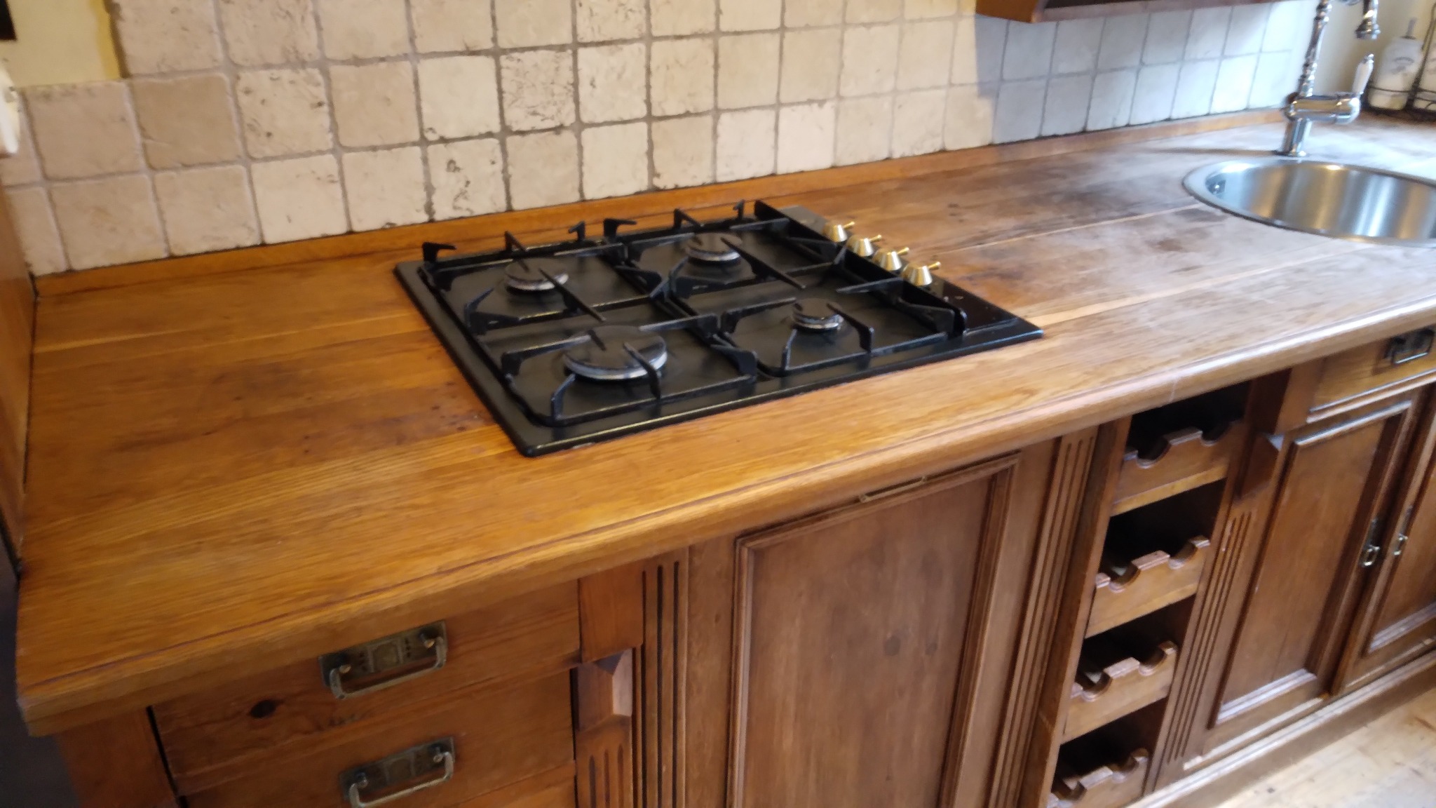 Drewniany blat kuchenny z wbudowaną płytą gazową i zlewozmywakiem. Widoczne szafki z dekoracyjnymi frezowaniami i uchwytami. Płytki ceramiczne na ścianie.