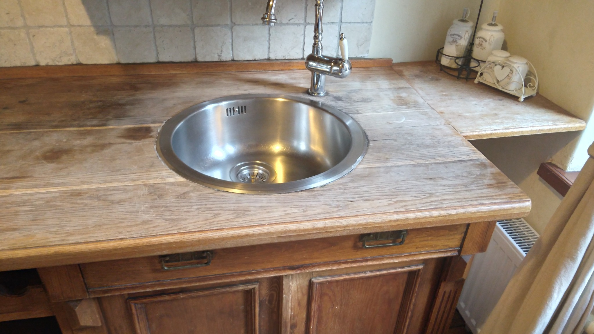 Rustykalny blat drewniany z wbudowanym zlewozmywakiem i baterią. Widoczne szafki pod blatem, dekoracje i płytki na ścianie w tle.