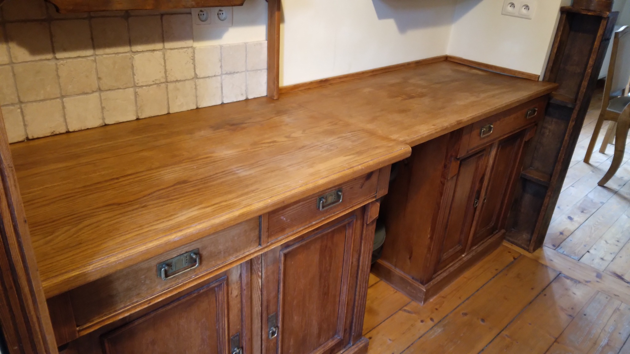 Rustykalna kuchnia z drewnianymi blatami i szafkami w ciepłym odcieniu brązu, widoczne płytki i drewniana podłoga. Styl retro z metalowymi uchwytami.