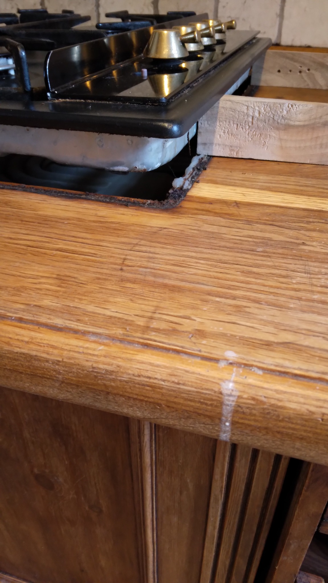 Fragment drewnianego blatu kuchennego z wyciętym otworem na płytę gazową. Widoczne niedoskonałości krawędzi i fragment szafki pod blatem.