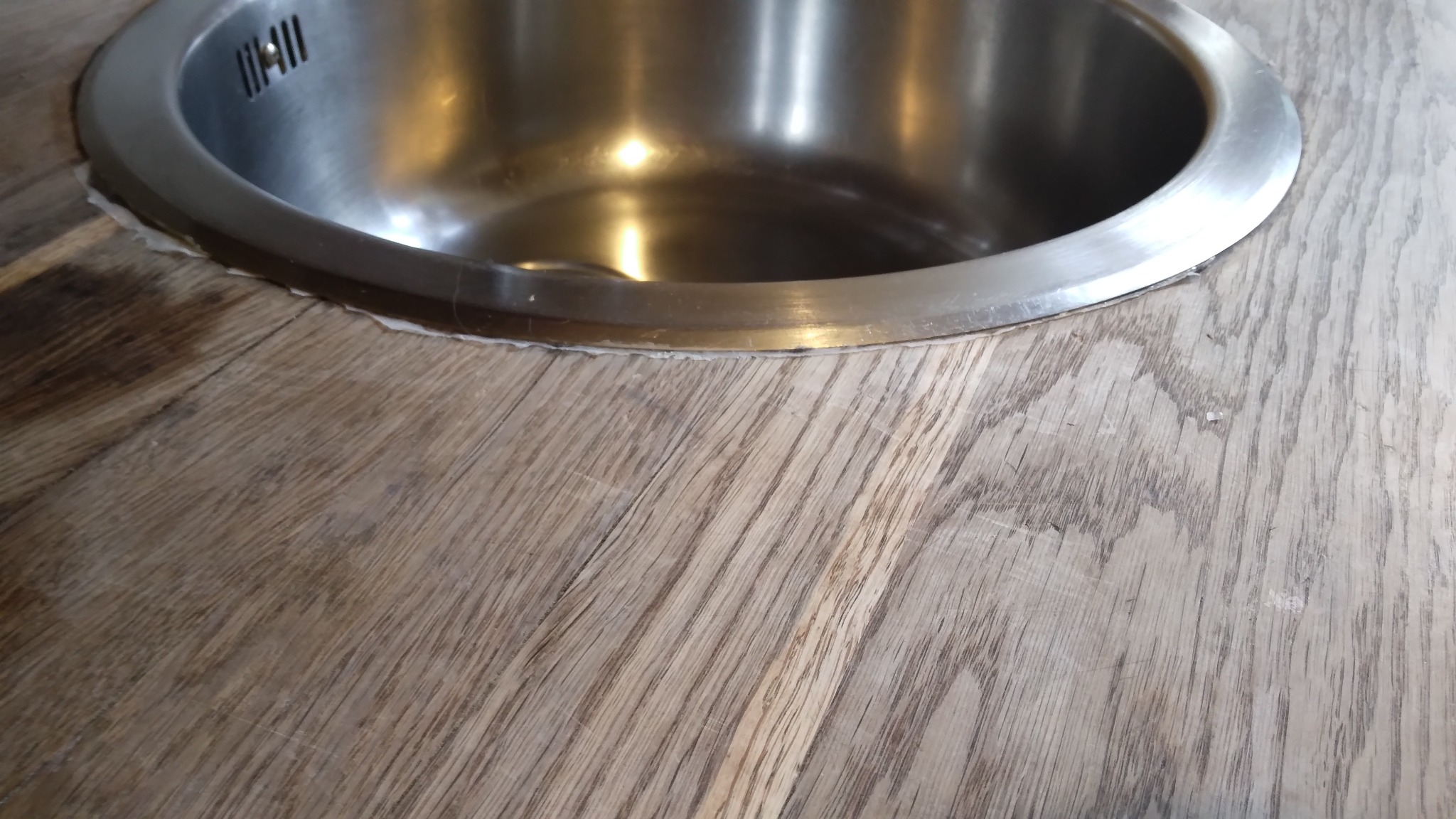 Okrągły, metalowy zlew wpuszczony w blat z jasnego drewna o wyraźnym usłojeniu. Widoczne delikatne niedoskonałości wykończenia wokół zlewu.