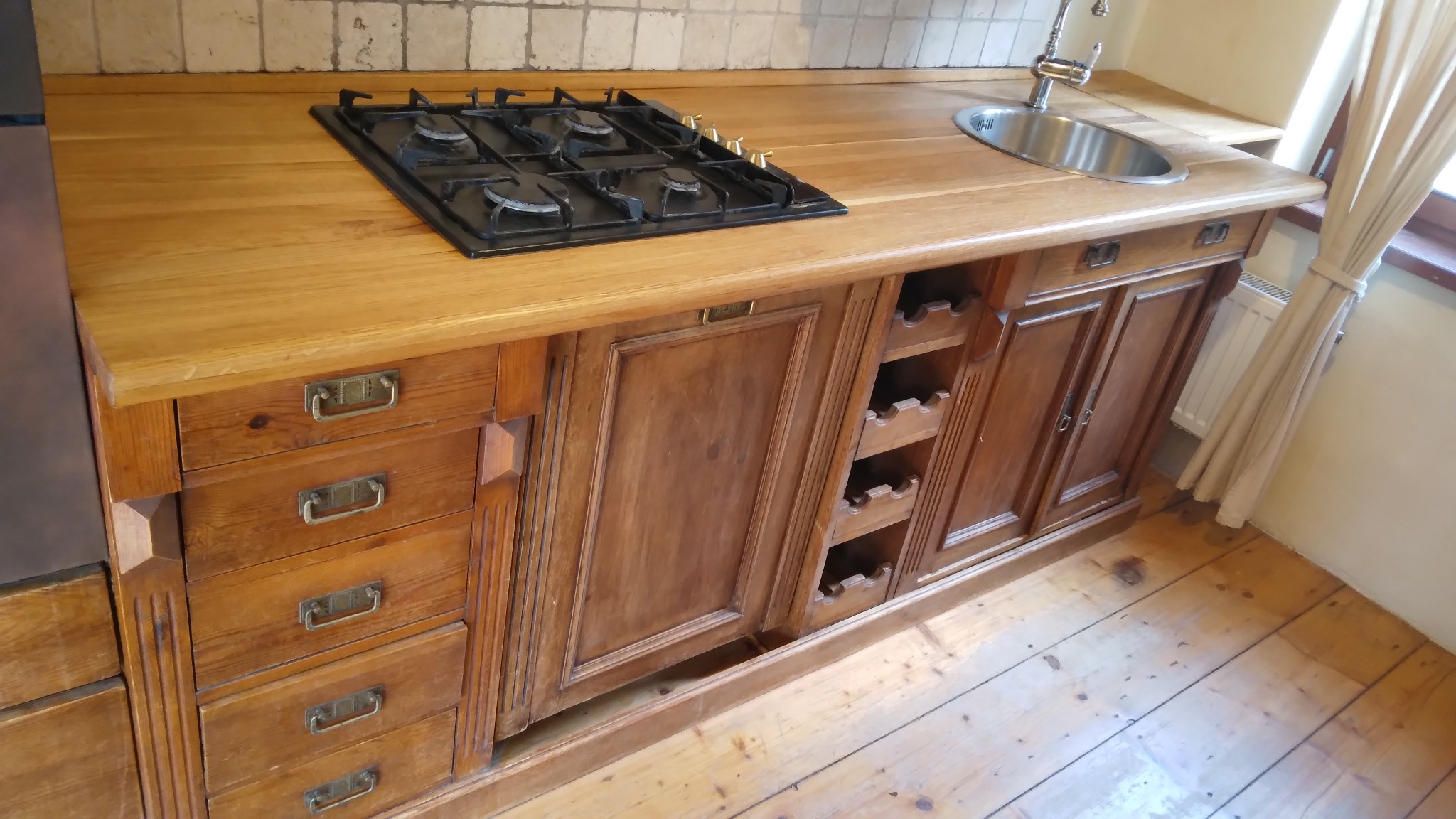 Rustykalna zabudowa kuchenna z drewnianym blatem, wbudowaną płytą grzewczą i zlewozmywakiem. Widoczne szuflady z metalowymi uchwytami i półki na wino.