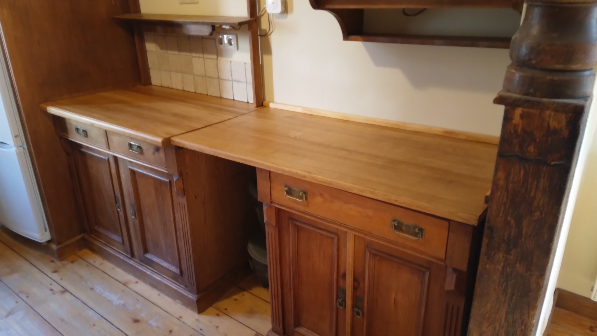 Rustykalna zabudowa kuchenna z drewnianymi szafkami, blatem i półkami. Widoczna jasna glazura nad blatem i drewniana podłoga. Charakterystyczny filar z ciemnego drewna.