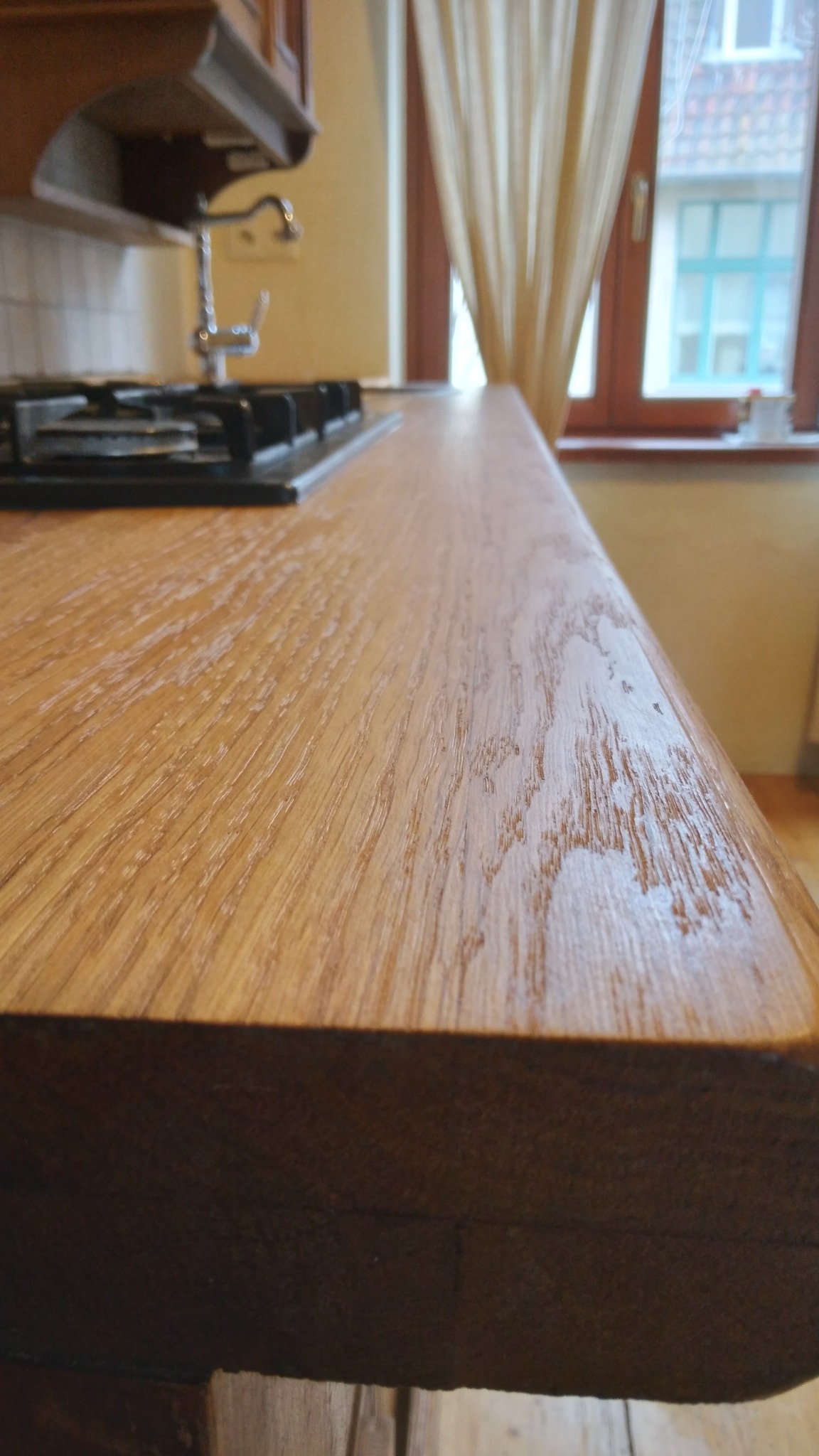 Drewniany blat kuchenny z zaokrąglonym brzegiem, widoczny fragment płyty grzewczej i okno w tle. Naturalne oświetlenie podkreśla strukturę drewna.