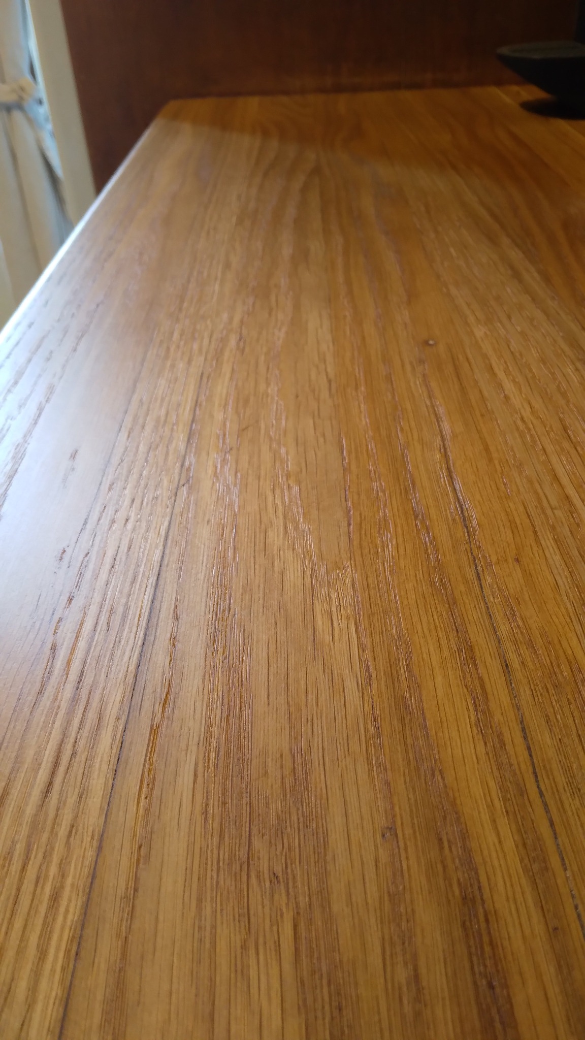 Jasny, lakierowany blat drewniany z wyraźnym usłojeniem, ukazany pod kątem. W tle fragment zasłony i ciemny element, prawdopodobnie monitora.