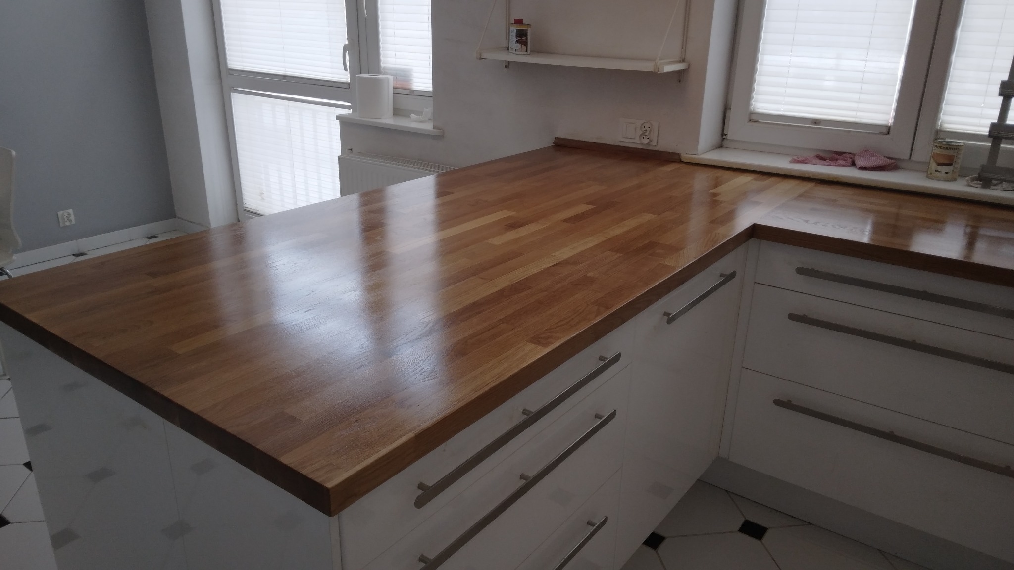 Polerowany blat drewniany w kuchni z białymi szafkami i srebrnymi uchwytami, pod oknem z żaluzjami. Blat odbija światło, ukazując unikalny wzór drewna.