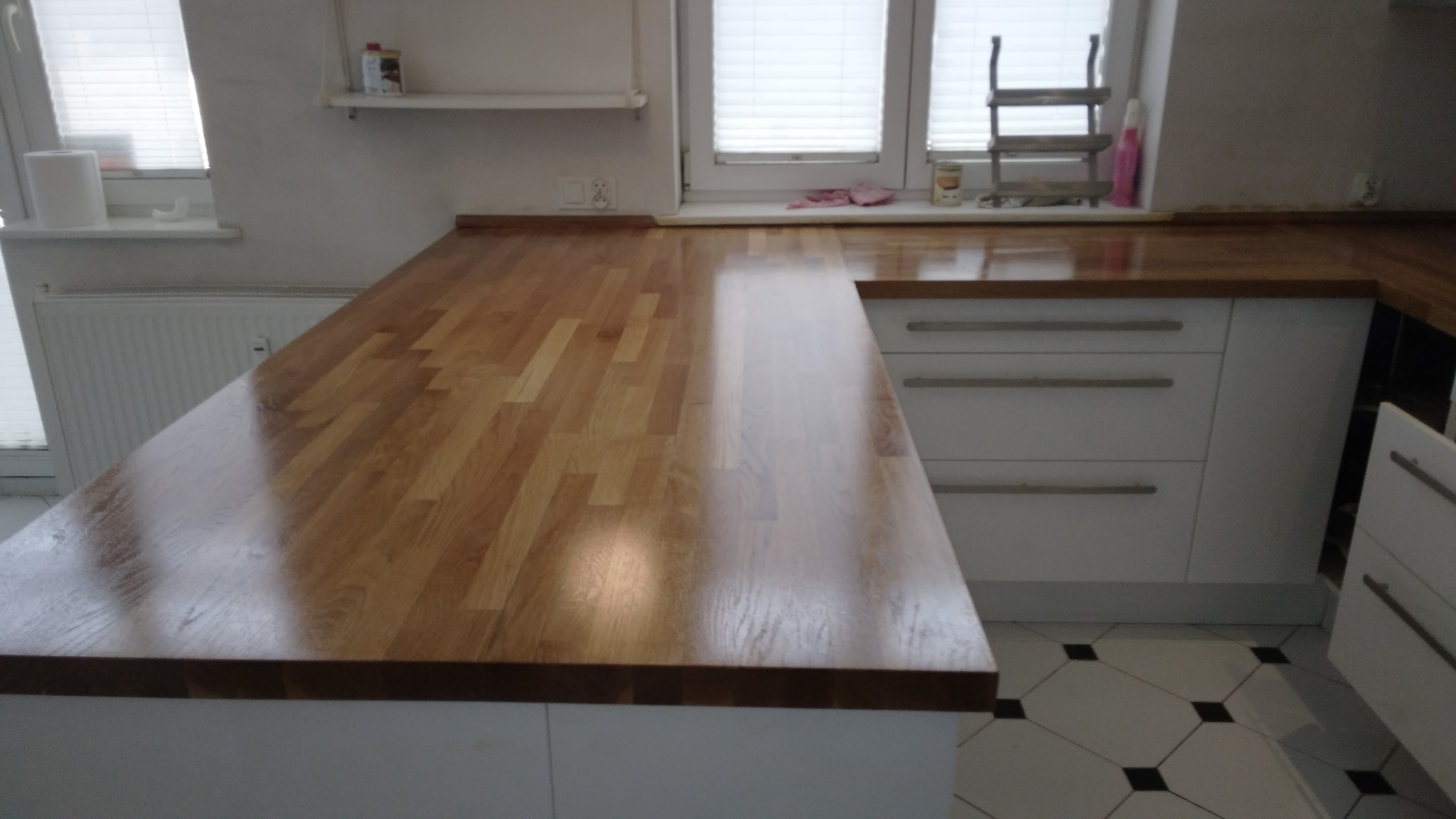 Jasny, drewniany blat kuchenny z widoczną strukturą drewna, zamontowany na białych szafkach. W tle okno z żaluzjami i akcesoria kuchenne.