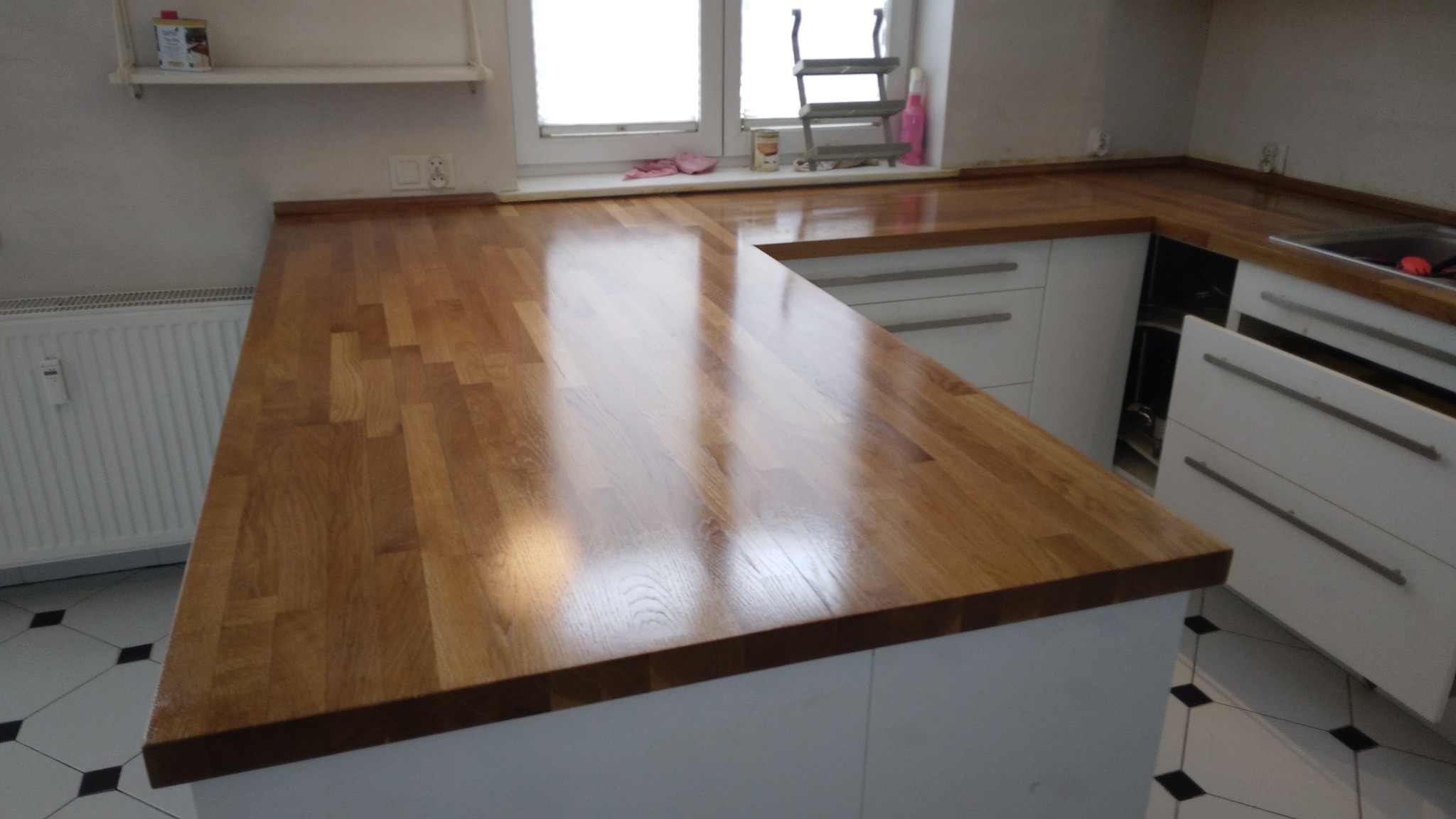 Kuchenny blat drewniany z widocznymi słojami, połączony z białymi szafkami, odbijający światło okna. Ujęcie z bliska eksponuje strukturę drewna i wykończenie.