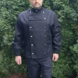 Mężczyzna w czarnym stroju kominiarskim i cylindrze pozuje na tle zielonego krzewu. Charakterystyczny ubiór i nakrycie głowy.