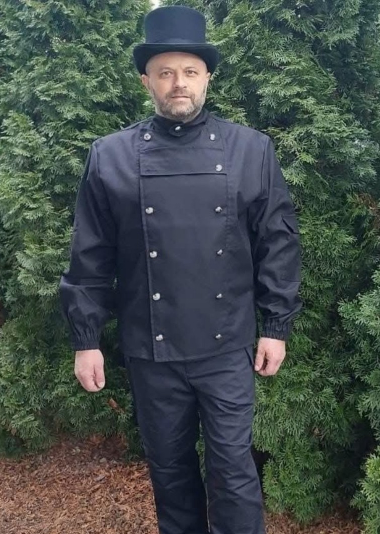 Mężczyzna w czarnym stroju kominiarskim i cylindrze pozuje na tle zielonego krzewu. Charakterystyczny ubiór i nakrycie głowy.