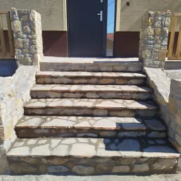 Kamienne schody wejściowe z drewnianą balustradą, prowadzące do ciemnych drzwi wejściowych.