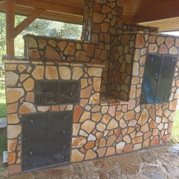 GIB-STONE MATEUSZ GIBEK - Zewnętrzny piec/grill z kamienia naturalnego z czarnymi, metalowymi drzwiczkami pod zadaszeniem, widok na zielony ogród w tle.