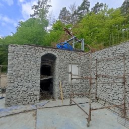 GIB-STONE MATEUSZ GIBEK - Budowa kamiennej konstrukcji z łukowym wejściem, z rusztowaniem i niebieskimi rurami na górze, w otoczeniu zieleni i drzew.