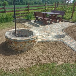 GIB-STONE MATEUSZ GIBEK - Ogród z kamiennym paleniskiem z rusztem, ścieżką z kamiennej kostki i drewnianym zestawem mebli ogrodowych na tle zieleni i drewnianego płotu.