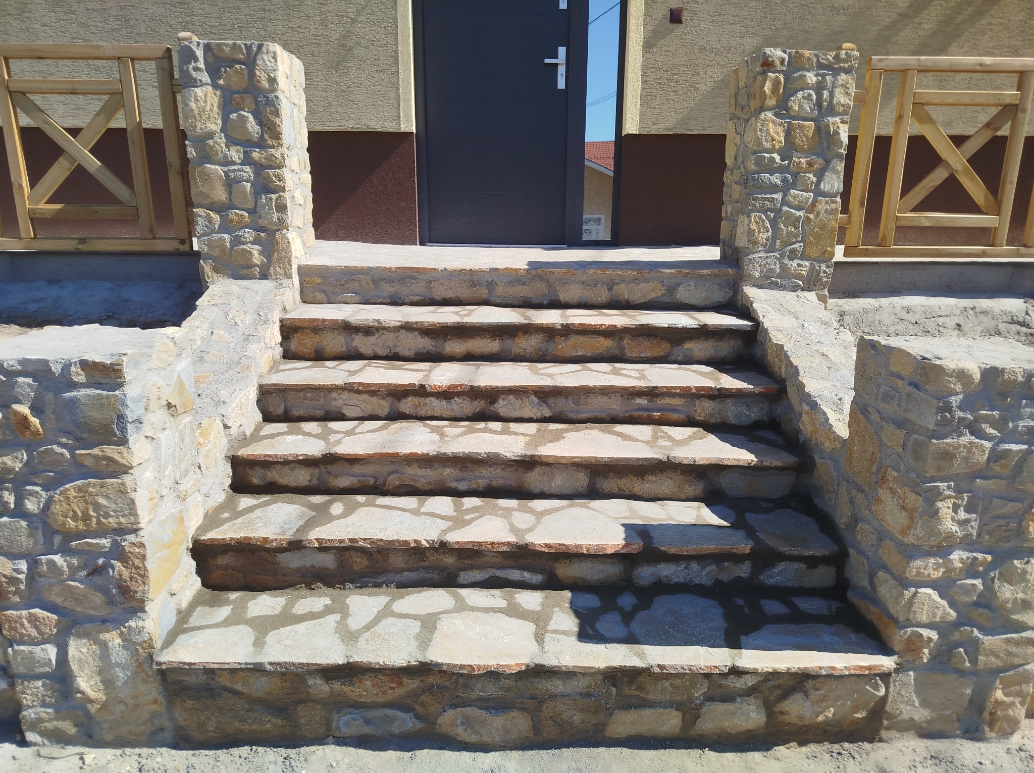 Kamienne schody wejściowe z drewnianą balustradą, prowadzące do ciemnych drzwi wejściowych.