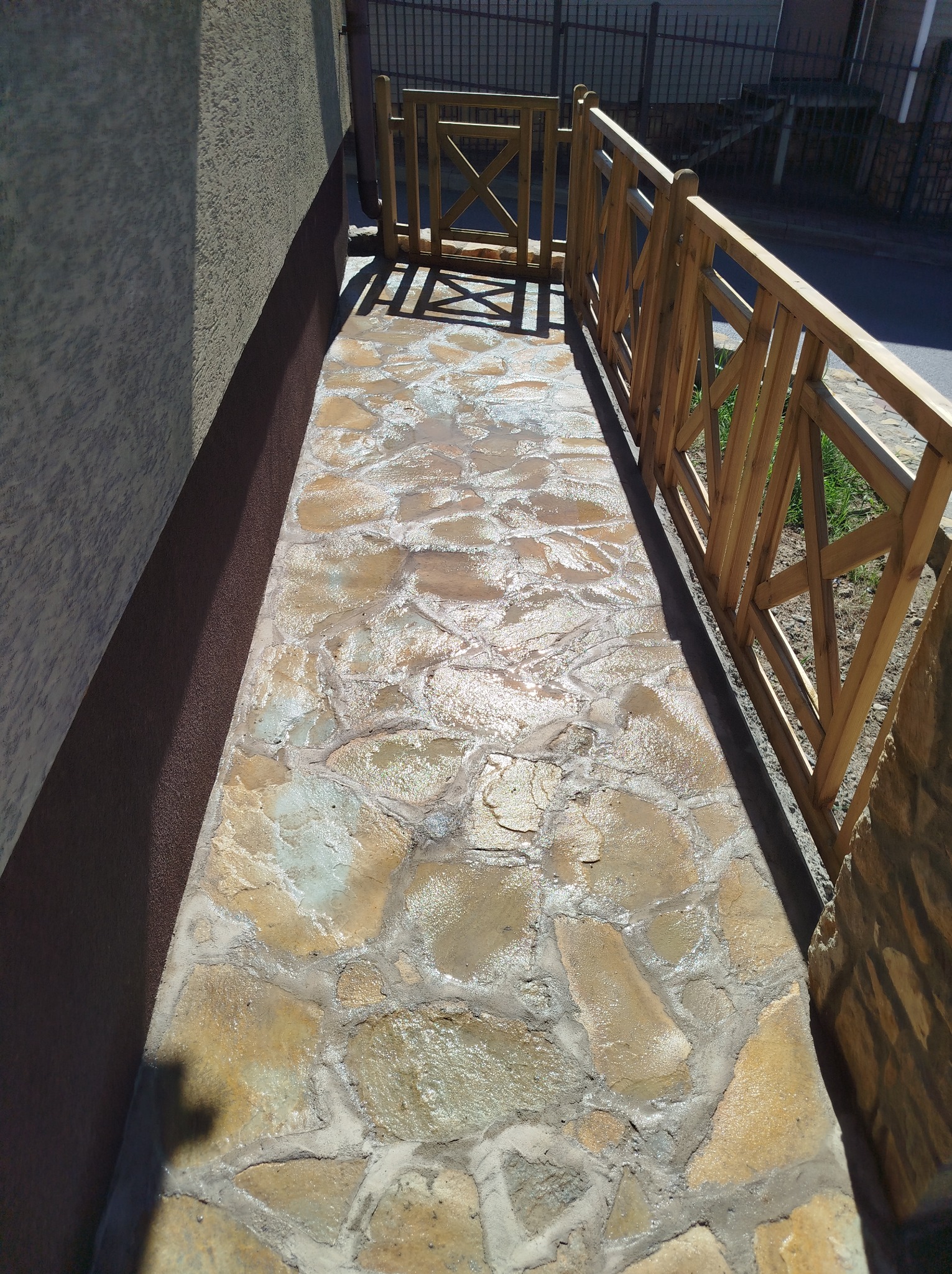 Ścieżka z nieregularnych kamieni, ograniczona drewnianym płotkiem, prowadząca do furtki. Widok z góry, w słoneczny dzień.