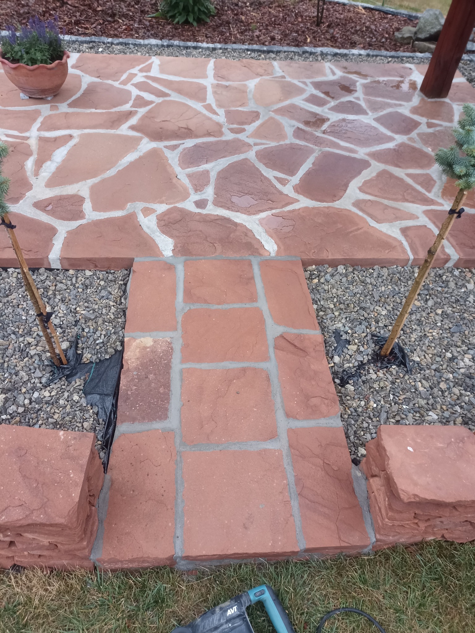 Czerwony taras z nieregularnych płyt kamiennych przechodzący w ścieżkę z kostki, obrzeża z kamienia, podsypane żwirem, widoczny fragment narzędzia