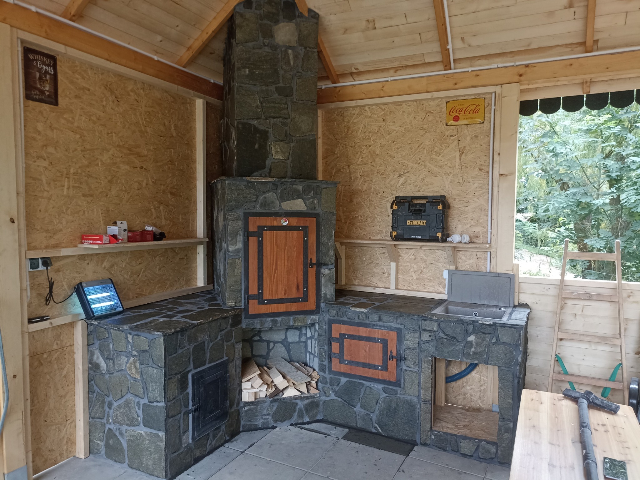 Kamienny grill z wędzarnią w drewnianej altanie, z drewnianymi drzwiczkami, ułożony stos drewna, widoczna skrzynka narzędziowa DeWalt, zlew i akcesoria