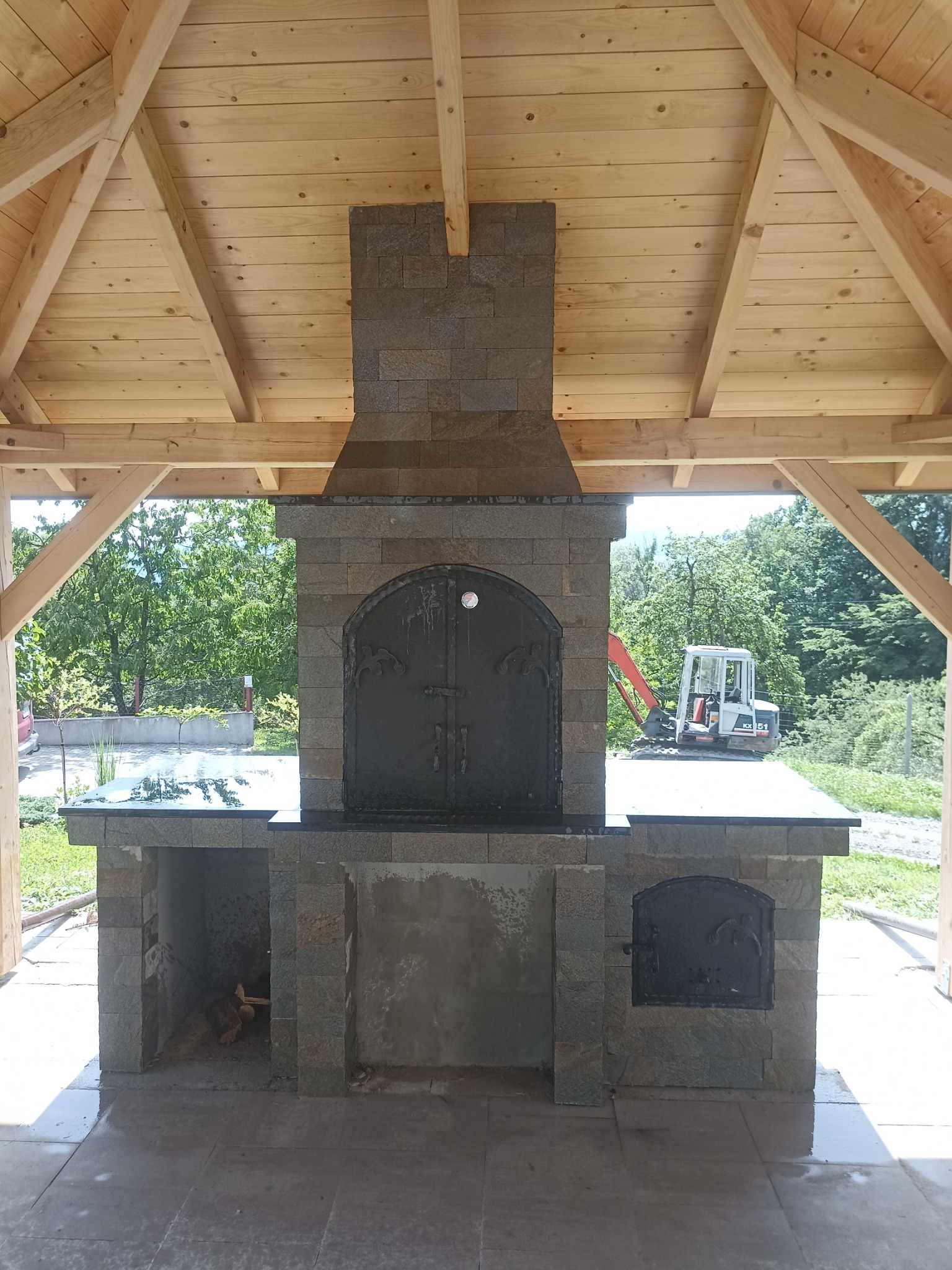 Kamienny grill z piekarnikiem pod drewnianą wiatą, z widoczną koparką w tle.