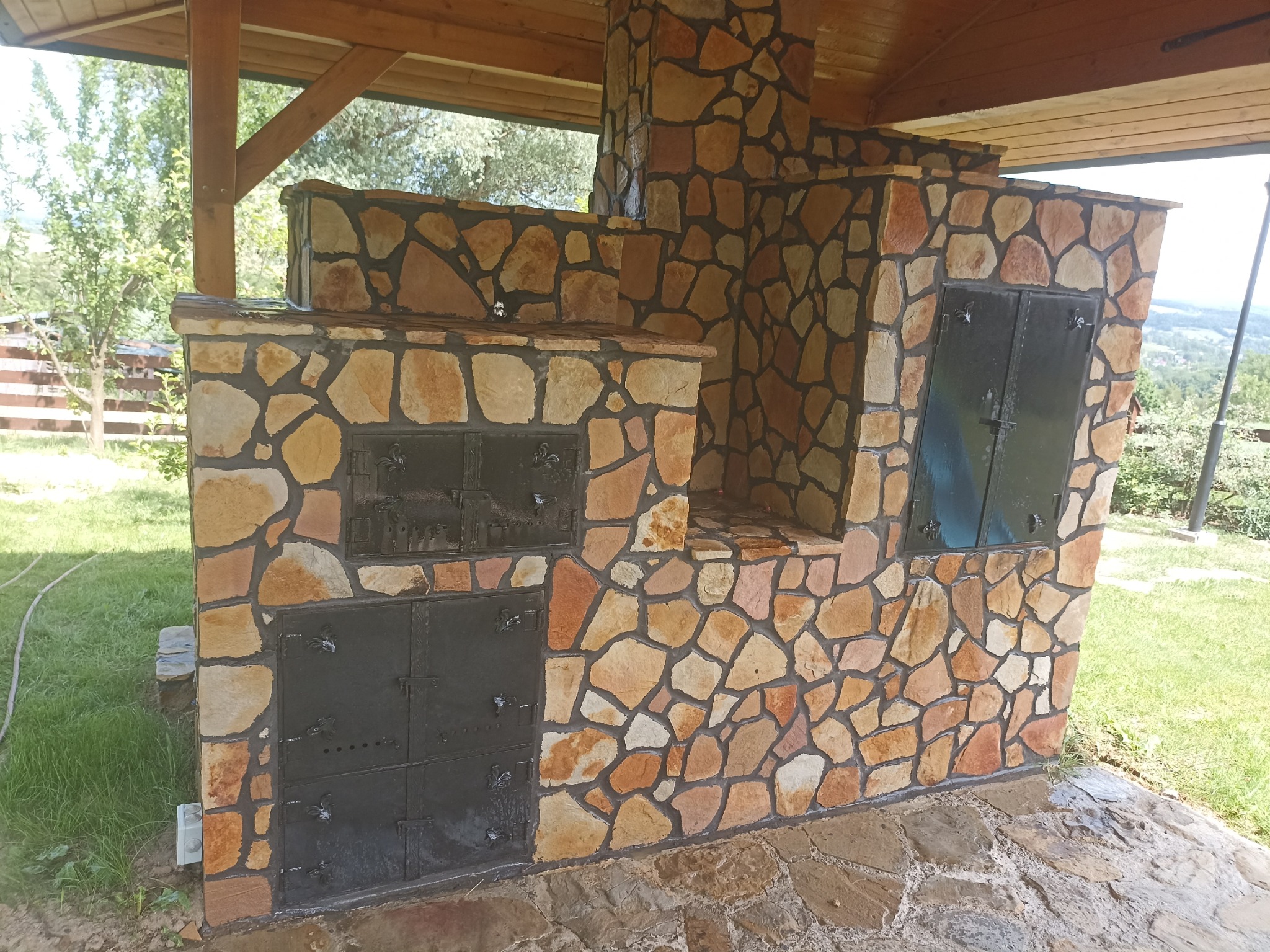 Zewnętrzny piec/grill z kamienia naturalnego z czarnymi, metalowymi drzwiczkami pod zadaszeniem, widok na zielony ogród w tle.