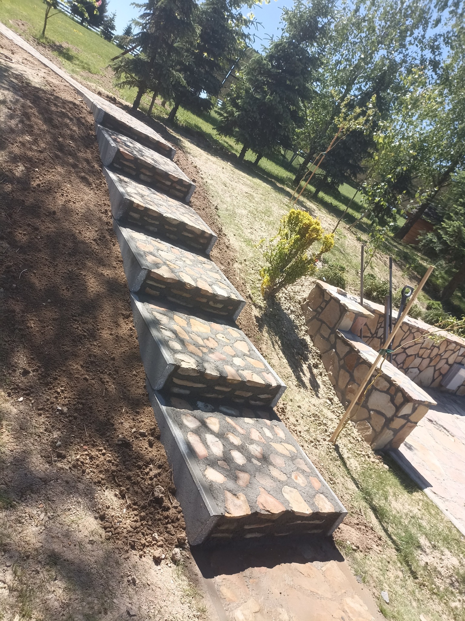 Kamienne schody ogrodowe z betonowymi obrzeżami w trakcie budowy, widoczne narzędzia i świeżo przekopana ziemia.