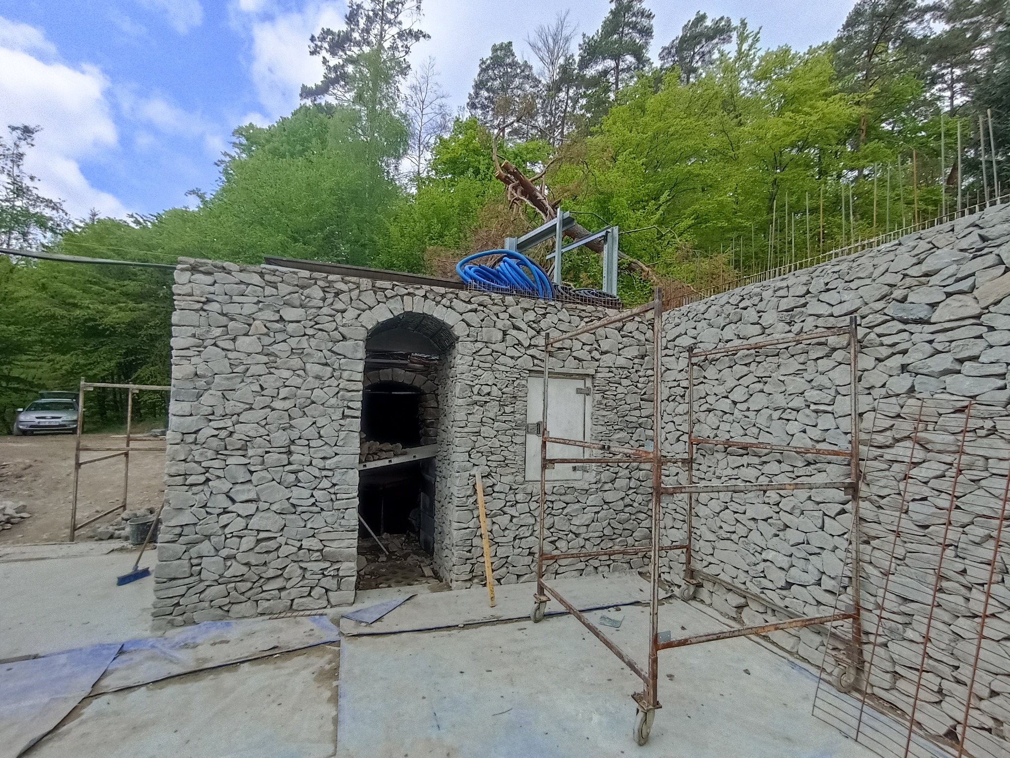 Budowa kamiennej konstrukcji z łukowym wejściem, z rusztowaniem i niebieskimi rurami na górze, w otoczeniu zieleni i drzew.