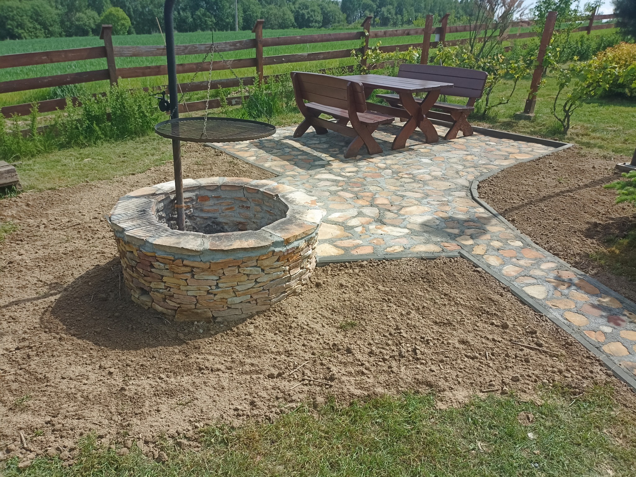 Ogród z kamiennym paleniskiem z rusztem, ścieżką z kamiennej kostki i drewnianym zestawem mebli ogrodowych na tle zieleni i drewnianego płotu.