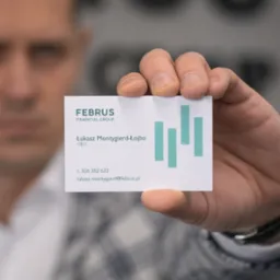 Mężczyzna w marynarce prezentuje wizytówkę firmy Februs Financial Group, z danymi kontaktowymi Łukasza Montygierd-Łojbo, CEO, na rozmytym tle biurowym.