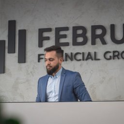 Februs Financial Group - Eksperci Kredytów Hipotecznych Łódź - Mężczyzna z brodą w niebieskiej marynarce stoi za biurkiem z logo 'FEBRUS FINANCIAL GROUP' na ścianie w tle.