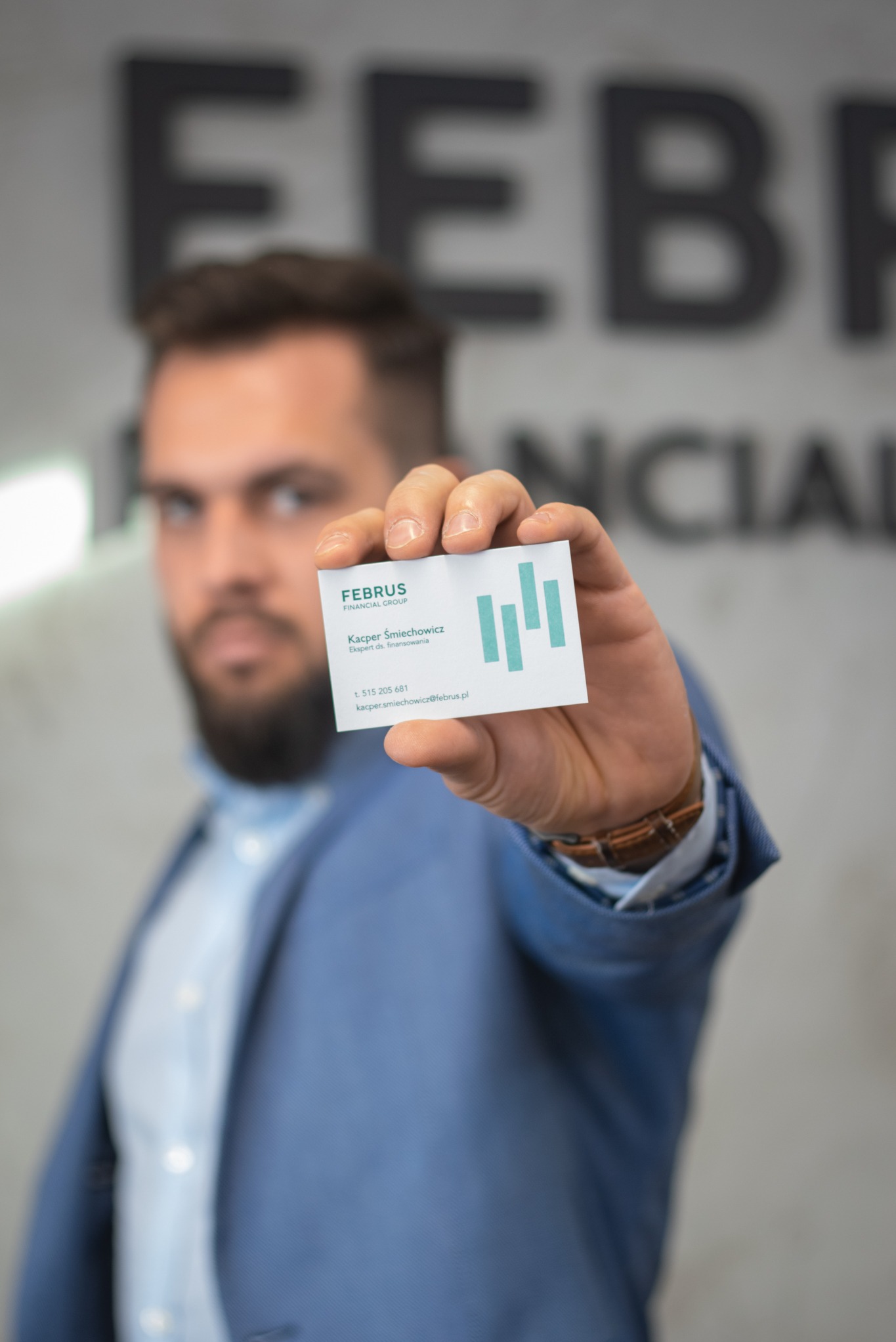 Mężczyzna w niebieskiej marynarce prezentuje wizytówkę firmy finansowej FEBRUS Financial Group, z danymi kontaktowymi Kacpra Śmiechowicza, eksperta ds. finansowania.