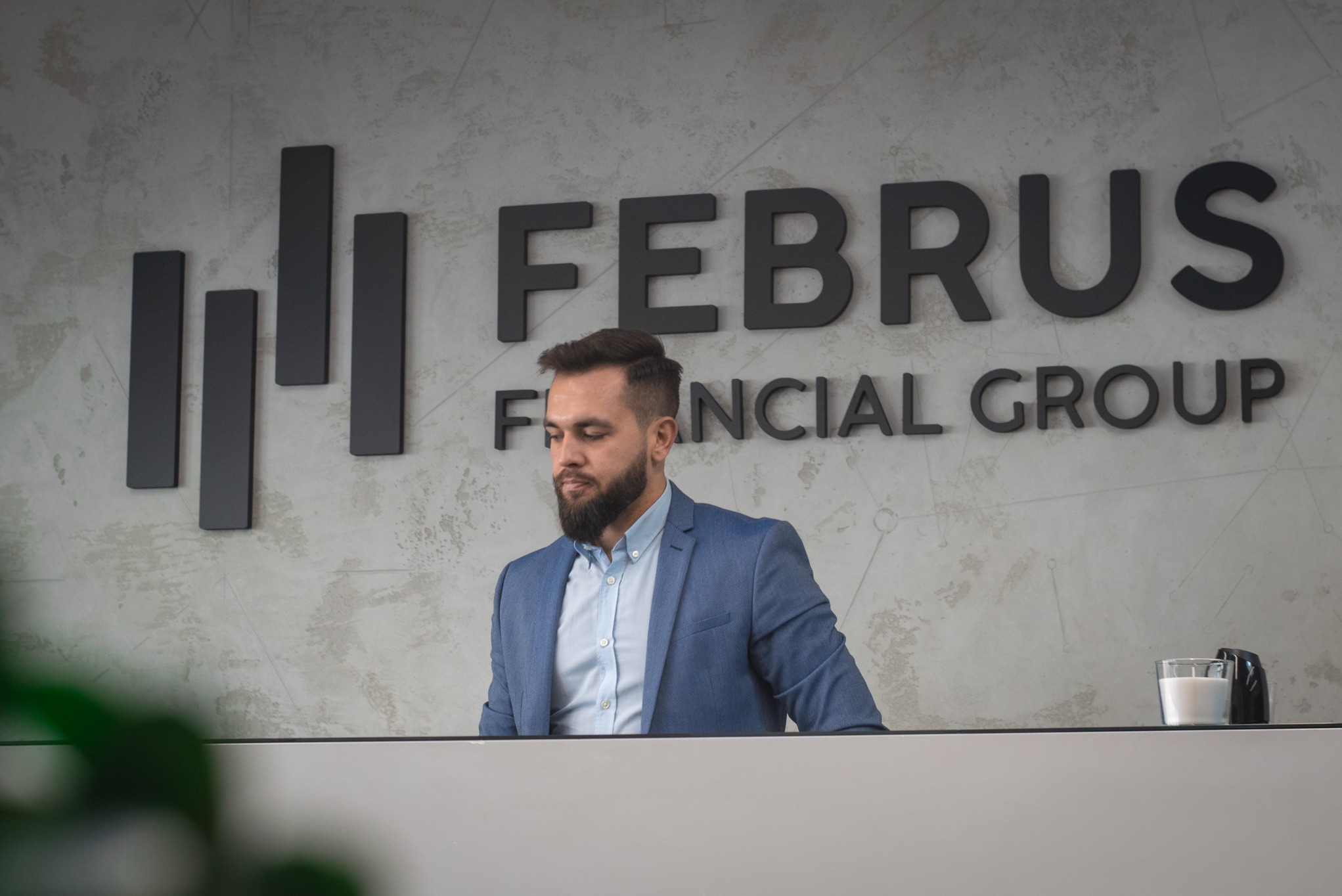 Mężczyzna z brodą w niebieskiej marynarce stoi za biurkiem z logo 'FEBRUS FINANCIAL GROUP' na ścianie w tle.
