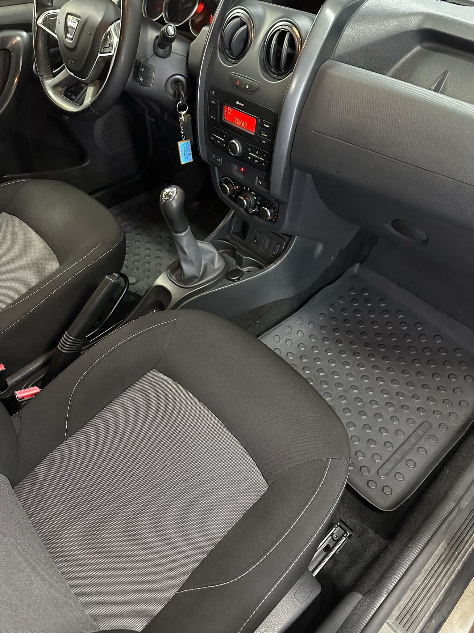 Wnętrze samochodu Dacia Duster, widoczne czarne i szare fotele z białymi przeszyciami, gumowe dywaniki z wytłoczonym wzorem, deska rozdzielcza z radiem i kluczykami w stacyjce, uchwyty na napoje.