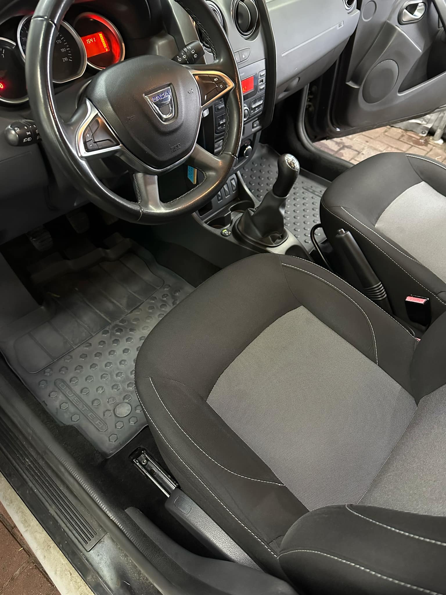 Wnętrze samochodu Dacia Duster: kierownica z logo, dwukolorowe fotele z jasnoszarą wstawką, gumowe dywaniki z wytłoczeniami.