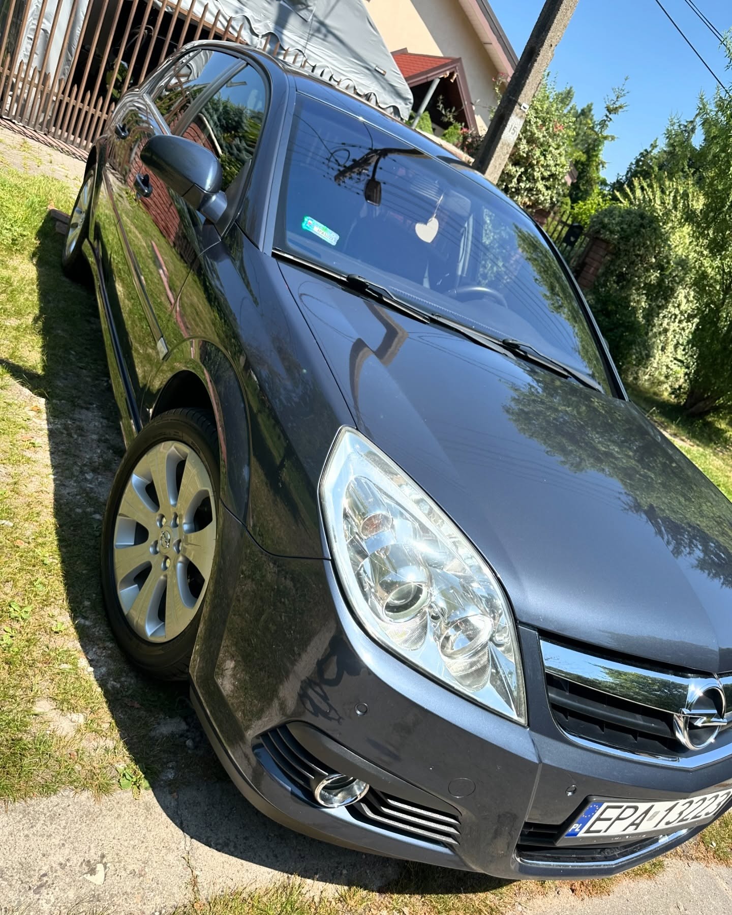 Szary samochód Opel Vectra stoi na trawie przed domem w słoneczny dzień, widoczny przód auta z reflektorami i tablicą rejestracyjną.