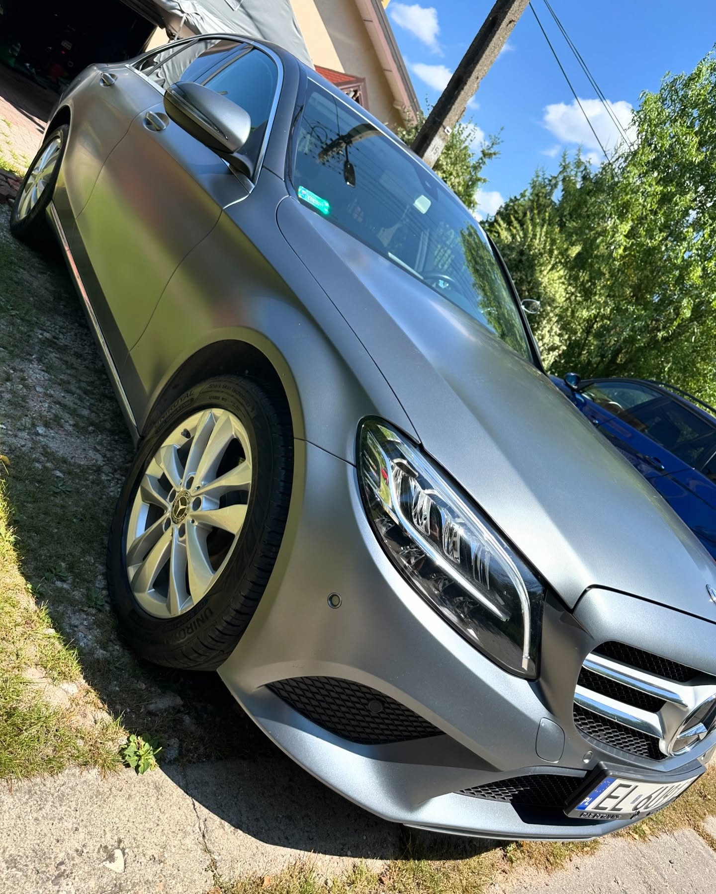 Szary Mercedes-Benz C-Class po zabiegu zmiany koloru folią matową, widoczny z bliska na tle zieleni i błękitnego nieba.