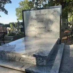 Jasnoszary, polerowany pomnik z granitu na cmentarzu, z wygrawerowanymi nazwiskami i datami, widoczne inne nagrobki i drzewa w tle.