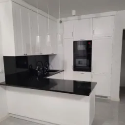 Nowoczesna kuchnia z białymi szafkami i czarnym blatem kamiennym z połyskiem, oświetlona trzema wiszącymi lampami cylindrycznymi.