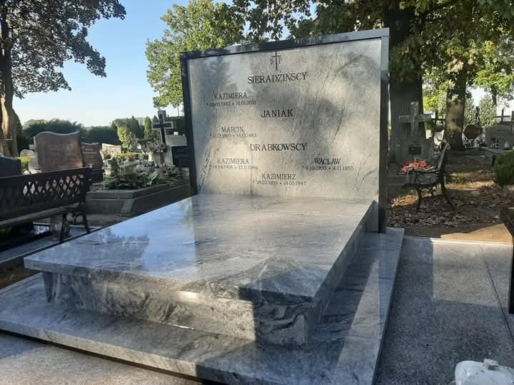 Jasnoszary, polerowany pomnik z granitu na cmentarzu, z wygrawerowanymi nazwiskami i datami, widoczne inne nagrobki i drzewa w tle.