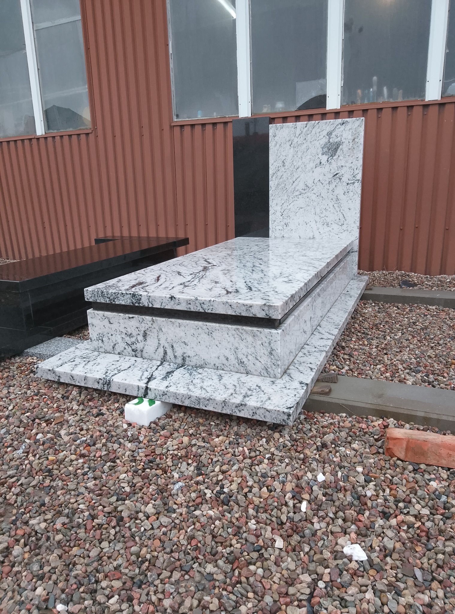 Gotowy nagrobek z jasnego, wzorzystego granitu z czarnym akcentem, ustawiony na żwirowym podłożu przed blaszanym budynkiem z oknami.