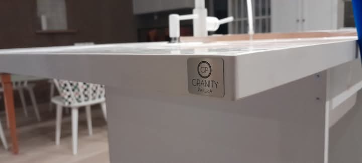 Biały blat kuchenny z logo 'Granity Pakuła' na krawędzi, umywalka i bateria w tle, widoczne krzesła i stół w pomieszczeniu.