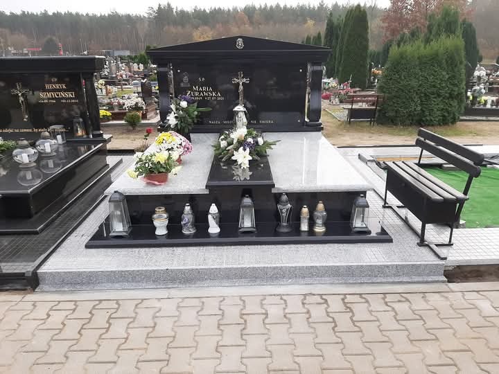 Czarny, granitowy pomnik nagrobny z jasnoszarym elementem centralnym, ozdobiony kwiatami i lampionami, umieszczony na cmentarzu z widocznymi innymi nagrobkami i ławką.