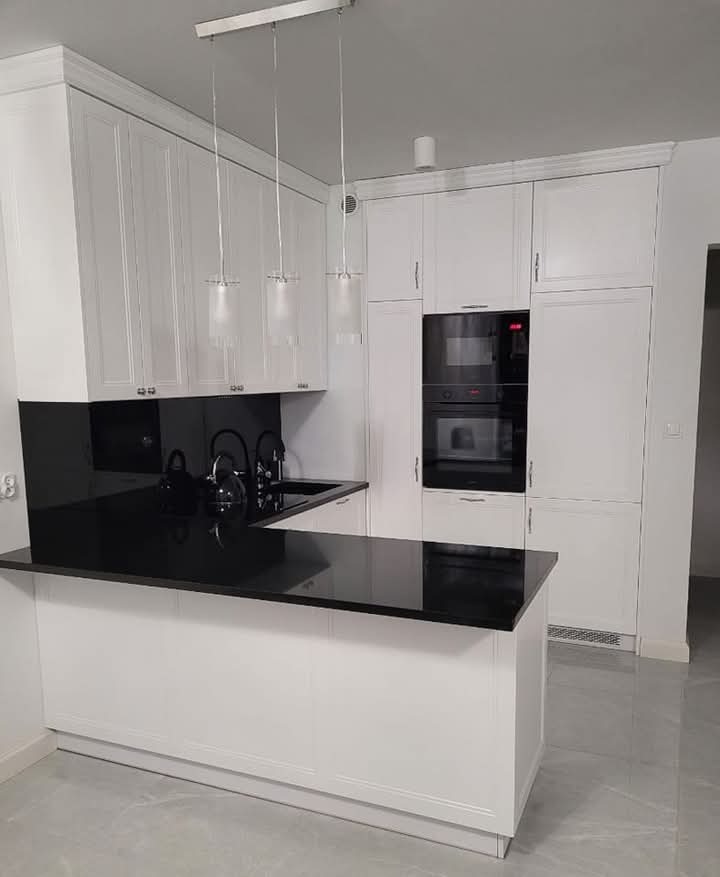 Nowoczesna kuchnia z białymi szafkami i czarnym blatem kamiennym z połyskiem, oświetlona trzema wiszącymi lampami cylindrycznymi.