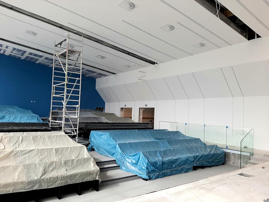 Sala konferencyjna w trakcie remontu: zabezpieczone folią krzesła, rusztowanie, sufit z widocznymi instalacjami, niebieska ściana w tle.