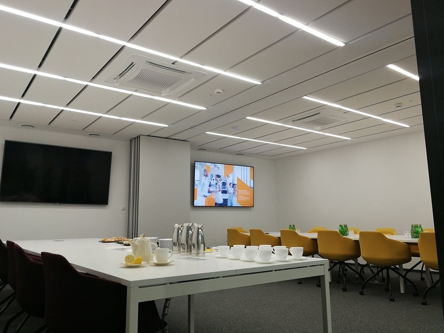 Nowoczesna sala konferencyjna z białym sufitem kasetonowym, liniowym oświetleniem LED, dwoma telewizorami, dużym białym stołem z zastawą i żółtymi krzesłami obrotowymi.